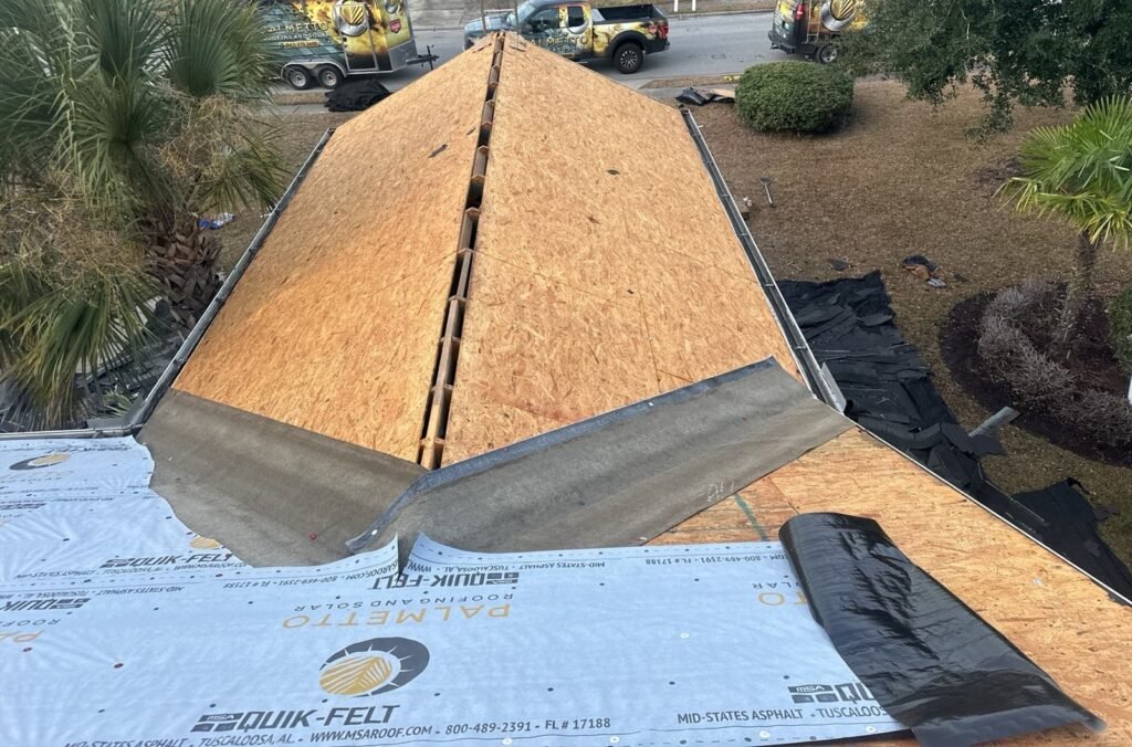 Overhead view of a hip roof stripped to decking with Palmetto Roofing branded truck, van and trailer at the curb