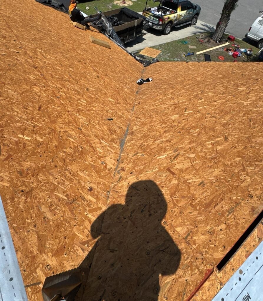 Roof stripped back to bare plywood decking with a Palmetto Roofing truck and dump trailer on the street below