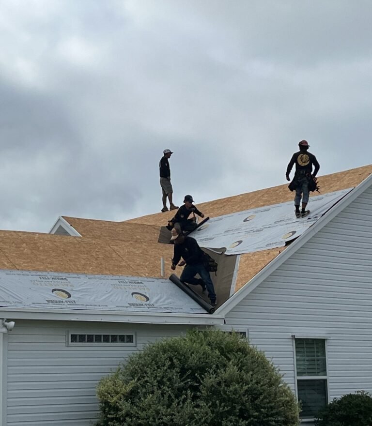 Four Palmetto roofers rolling synthetic underlayment across a stripped roof deck under a gray sky