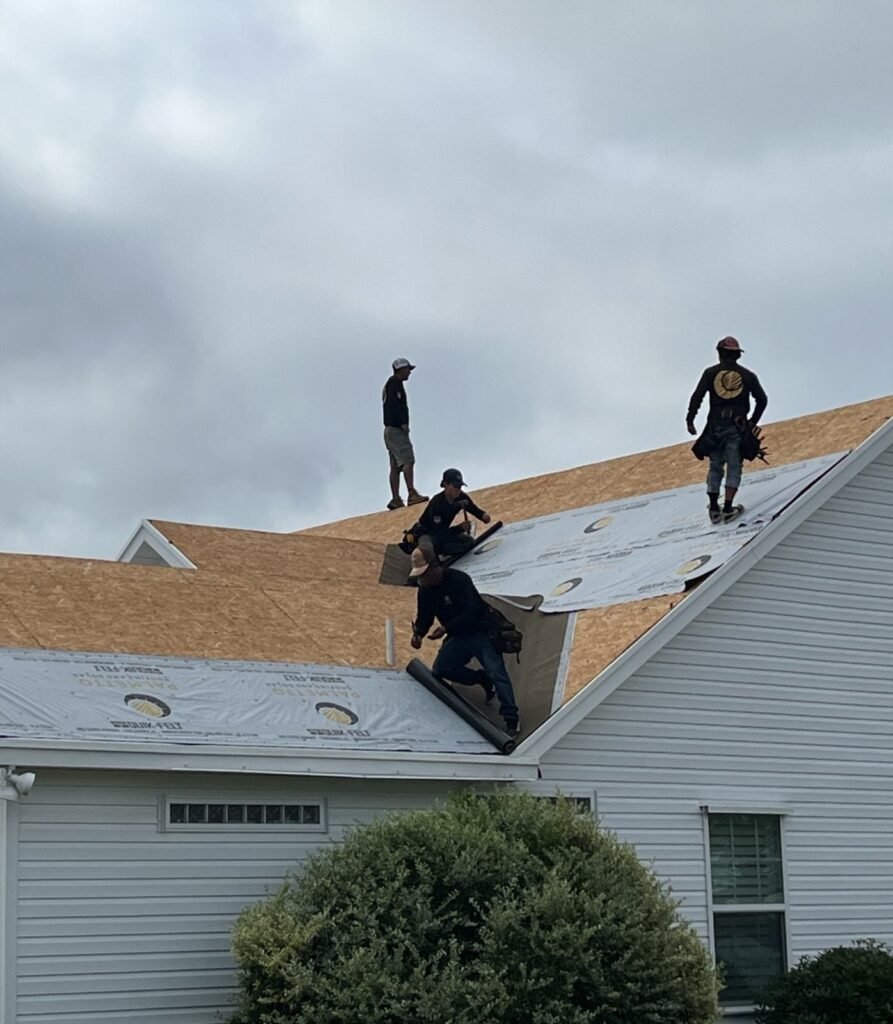 Four Palmetto roofers rolling synthetic underlayment across a stripped roof deck under a gray sky